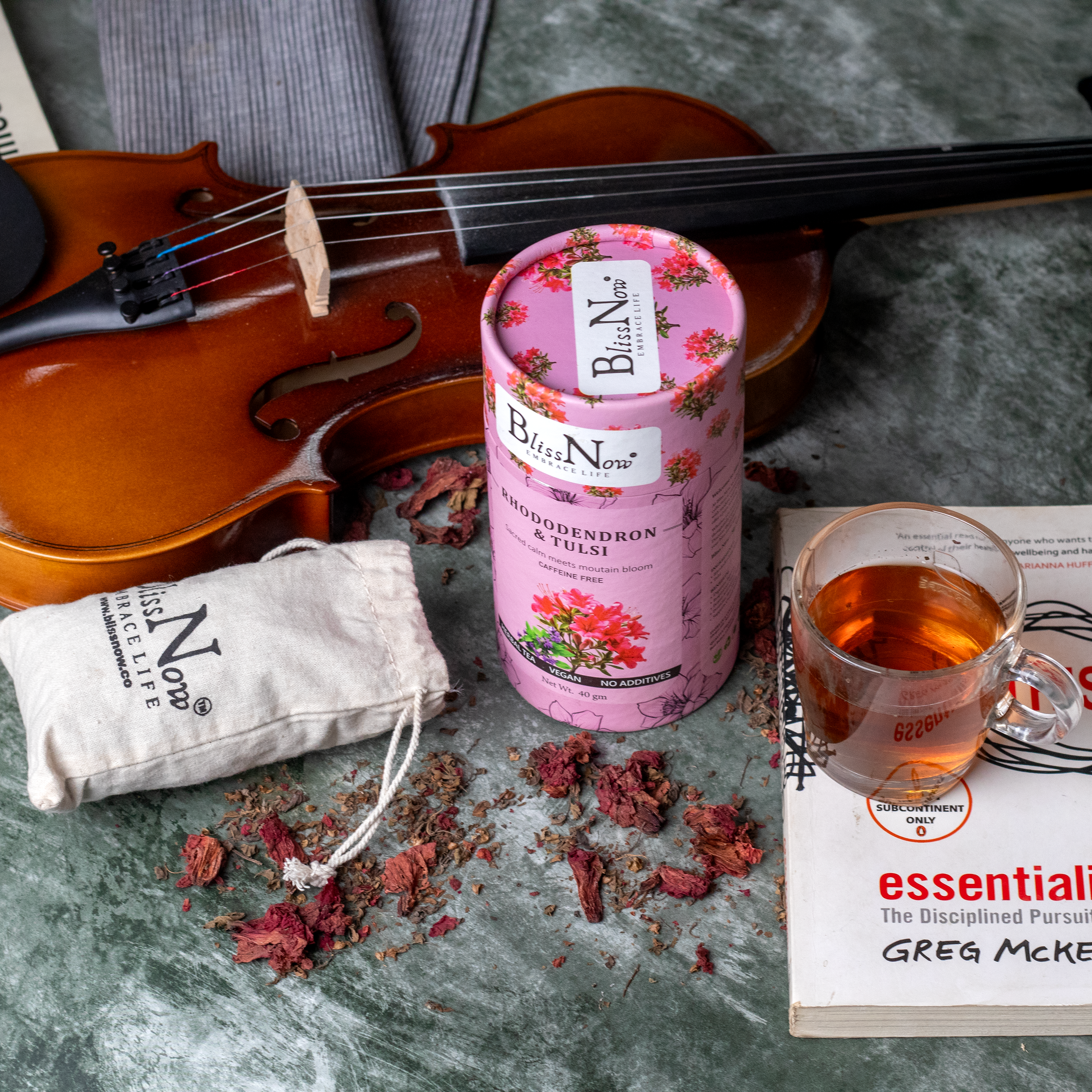 Bliss Now tea packaging with a cup of tea, dried flowers, and a violin on a textured surface.